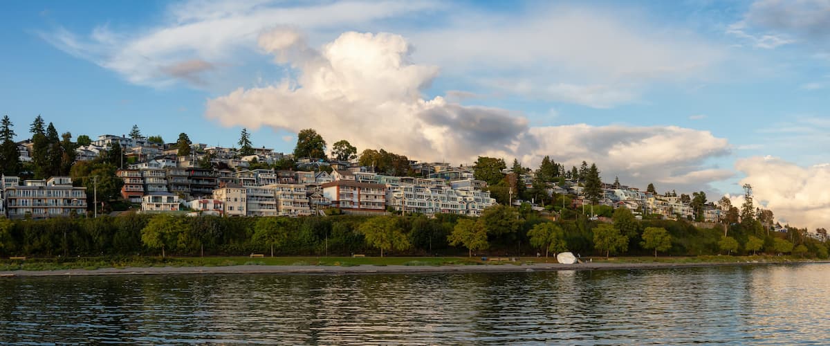 A waterfront cityscape with dense residential buildings, a calm river, and a backdrop of clouds.