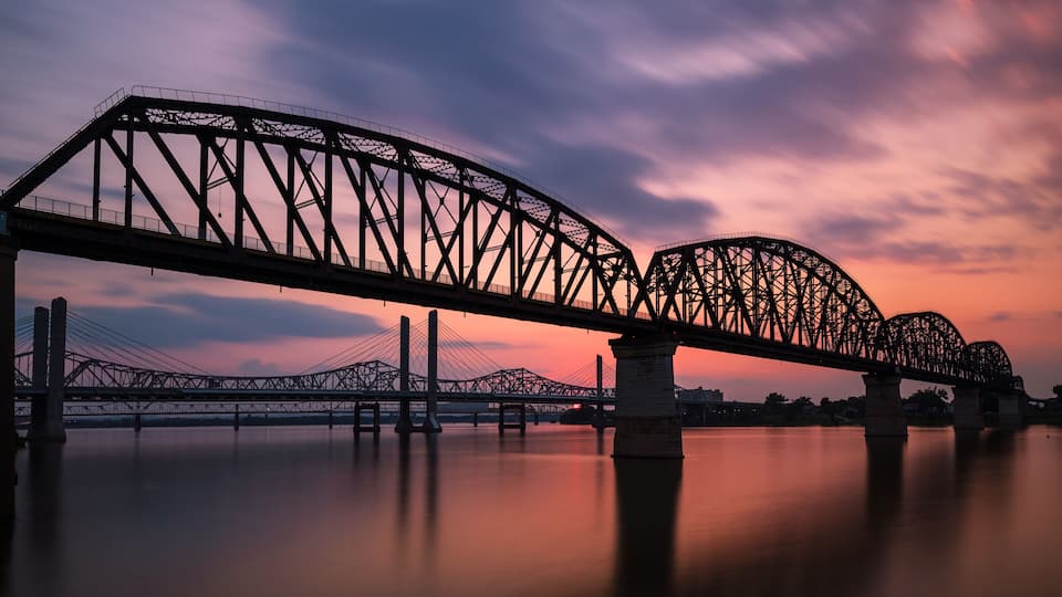 A steel truss bridge over calm water at sunset.