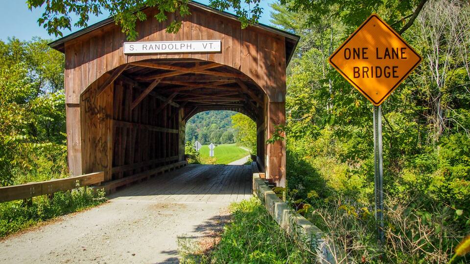 A wooden covered bridge with a sign reading "S. RANDOLPH, VT." and a "ONE LANE BRIDGE" sign.