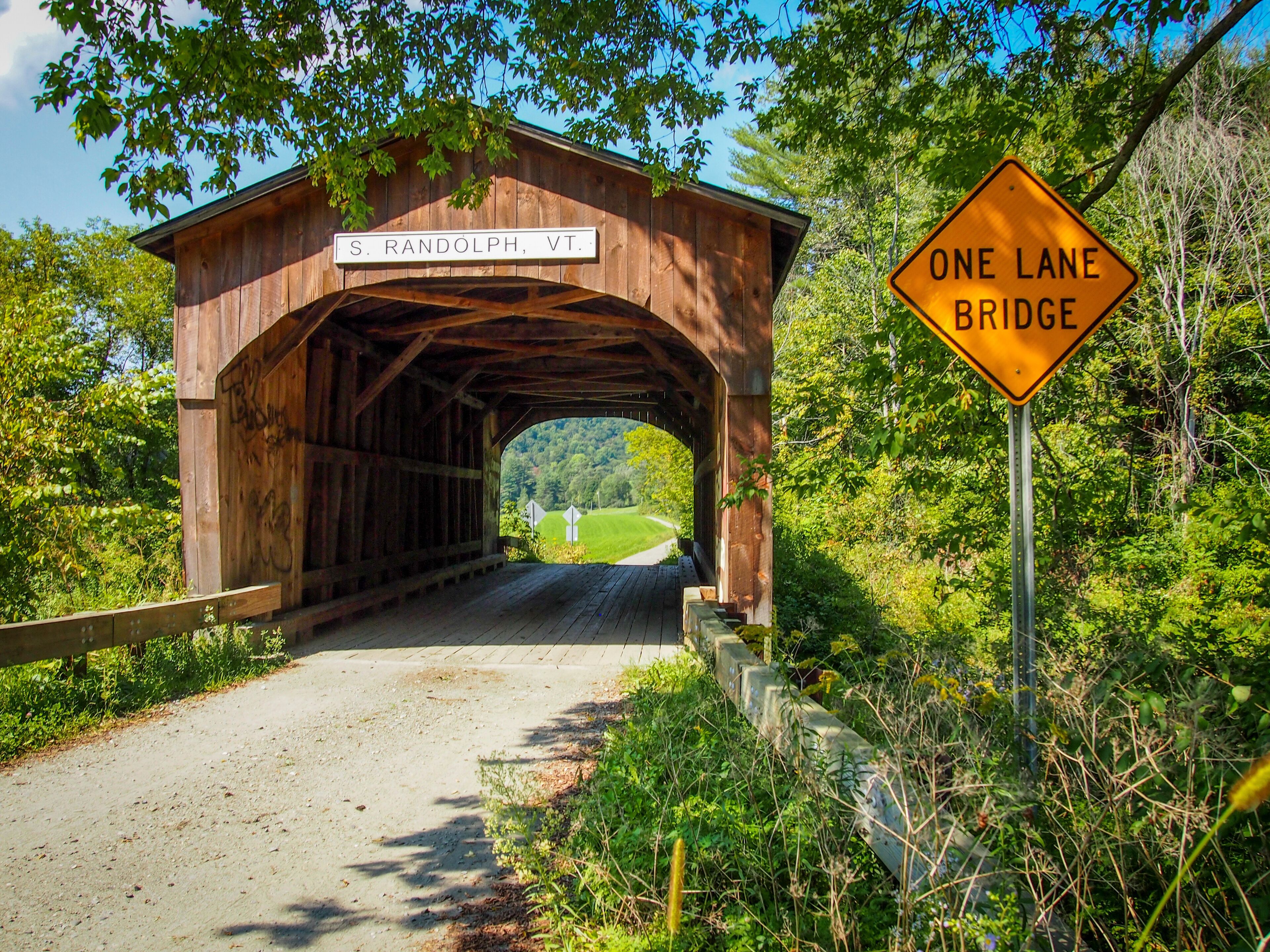 A wooden covered bridge with a sign reading "S. RANDOLPH, VT." and a "ONE LANE BRIDGE" sign.