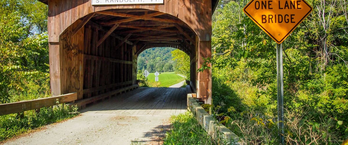 A wooden covered bridge with a sign reading "S. RANDOLPH, VT." and a "ONE LANE BRIDGE" sign.
