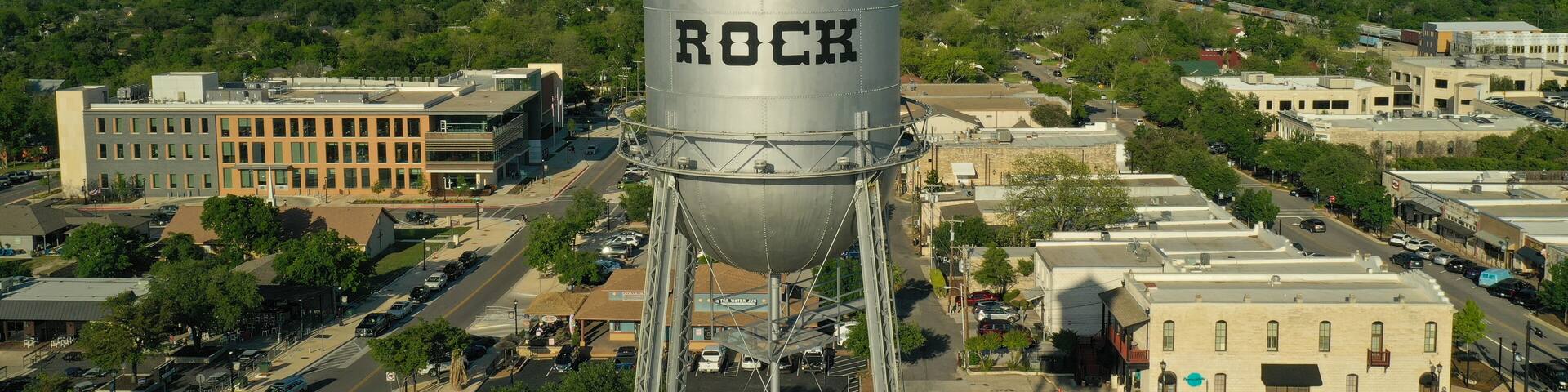 A water tower labeled "ROUND ROCK" with a clear blue sky and a cityscape in the background.