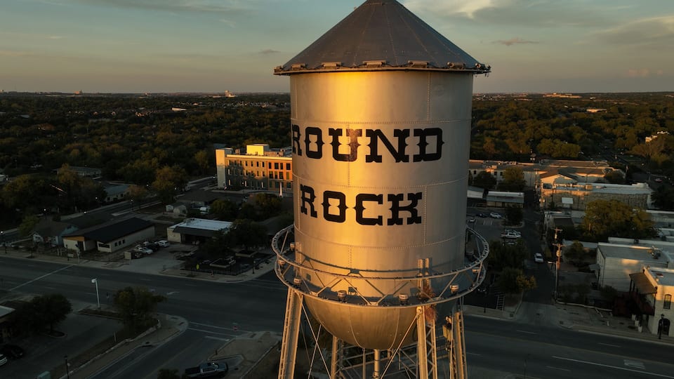 A water tower labeled 'ROUND ROCK' with a metal structure and a conical roof, surrounded by a cityscape with roads and buildings.
