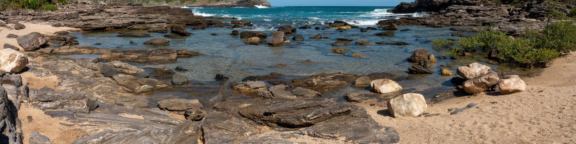 A rocky shoreline with clear blue water, a sandy beach, and green hills in the background.