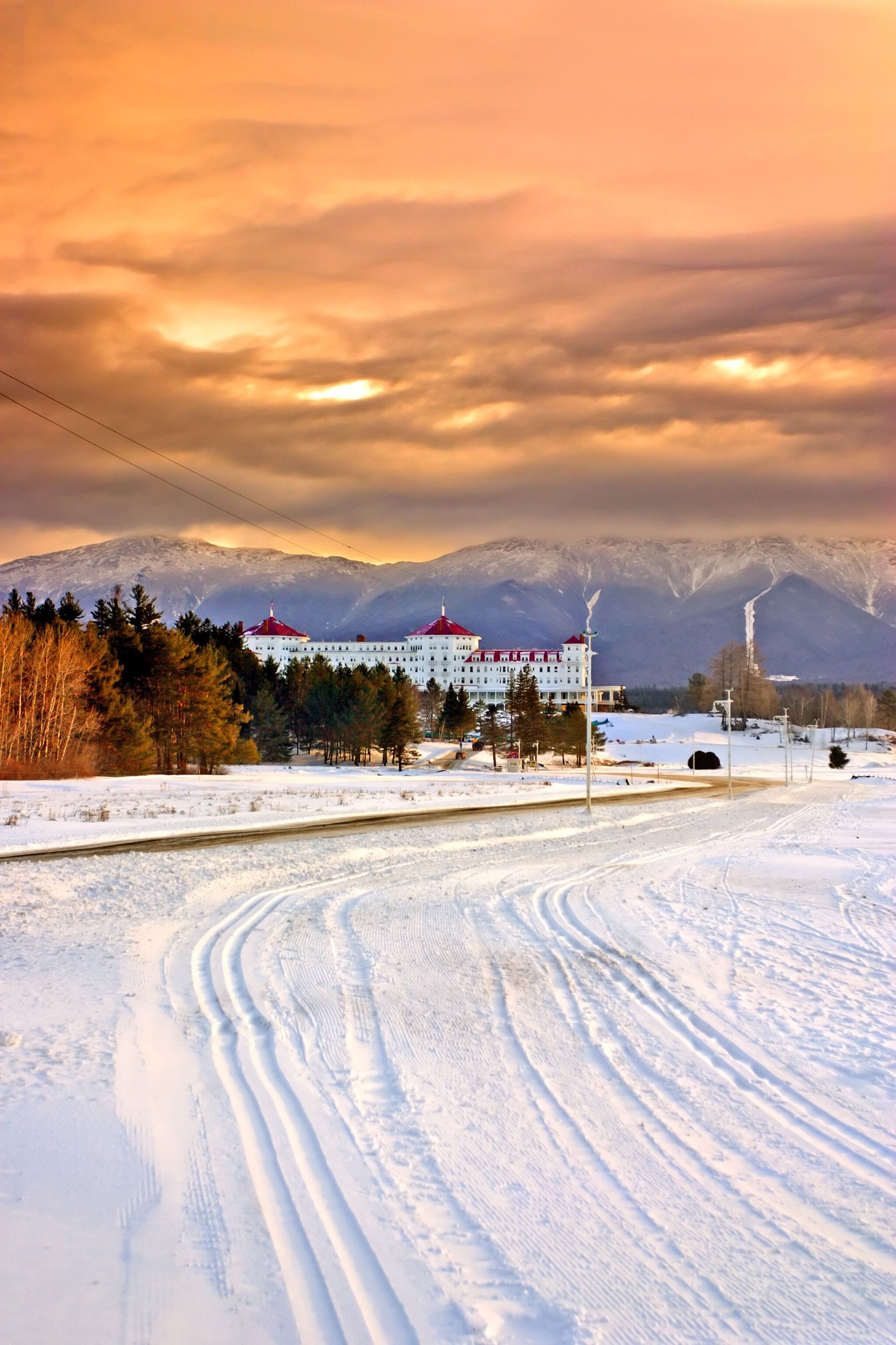 Paisagem coberta de neve com um edifício grande e telhados vermelhos, uma cordilheira ao fundo e céu claro.