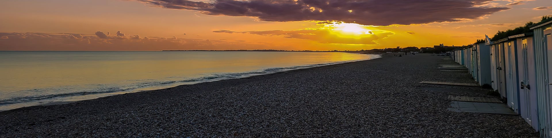 A pebbled beach with a row of seaside buildings and a sunset over the ocean.