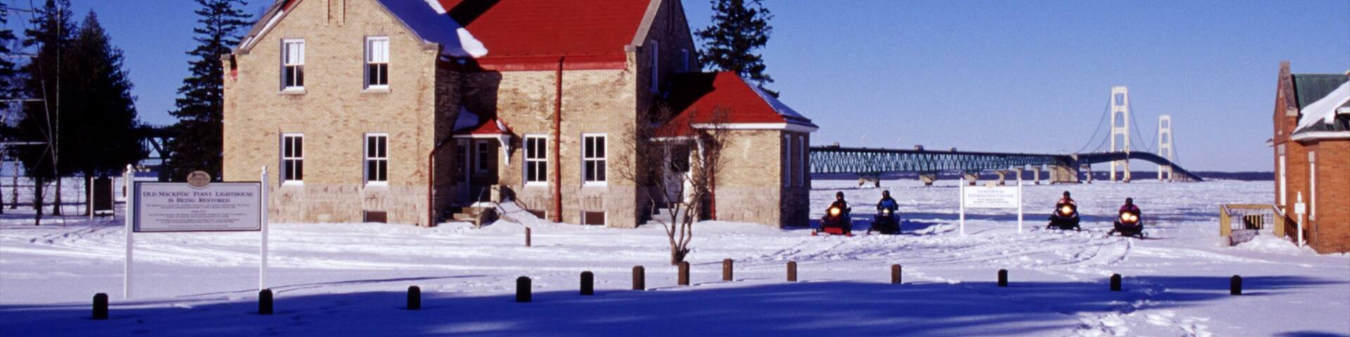 A historic lighthouse with a red roof, a bridge in the background, and snow-covered ground.