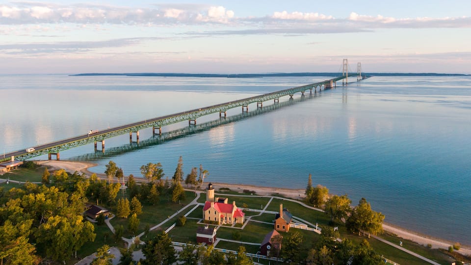 A long green bridge over a calm body of water, surrounded by trees and a small town with historic buildings.