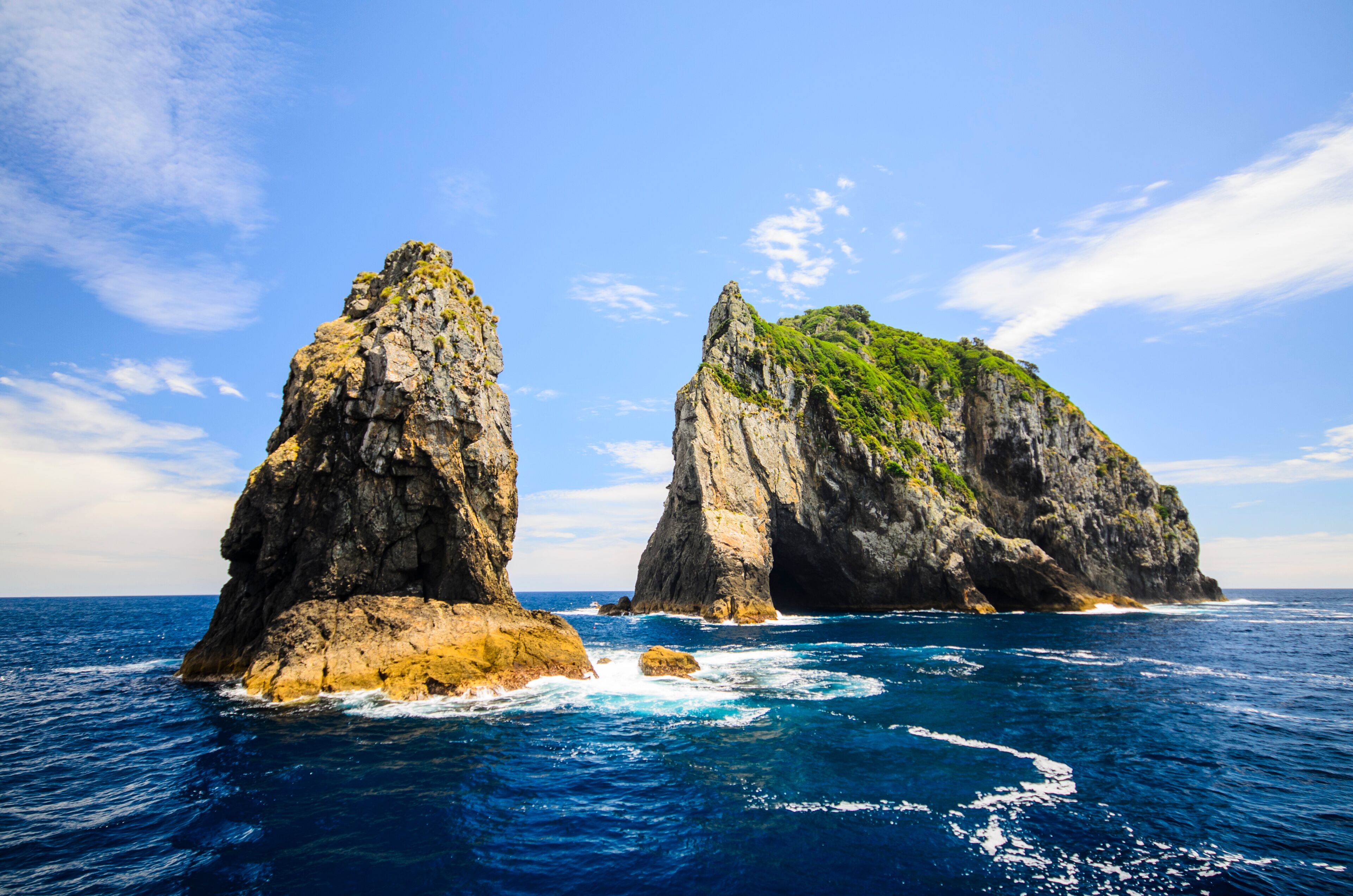 Two prominent rock formations in the ocean with clear blue water and a partly cloudy sky.