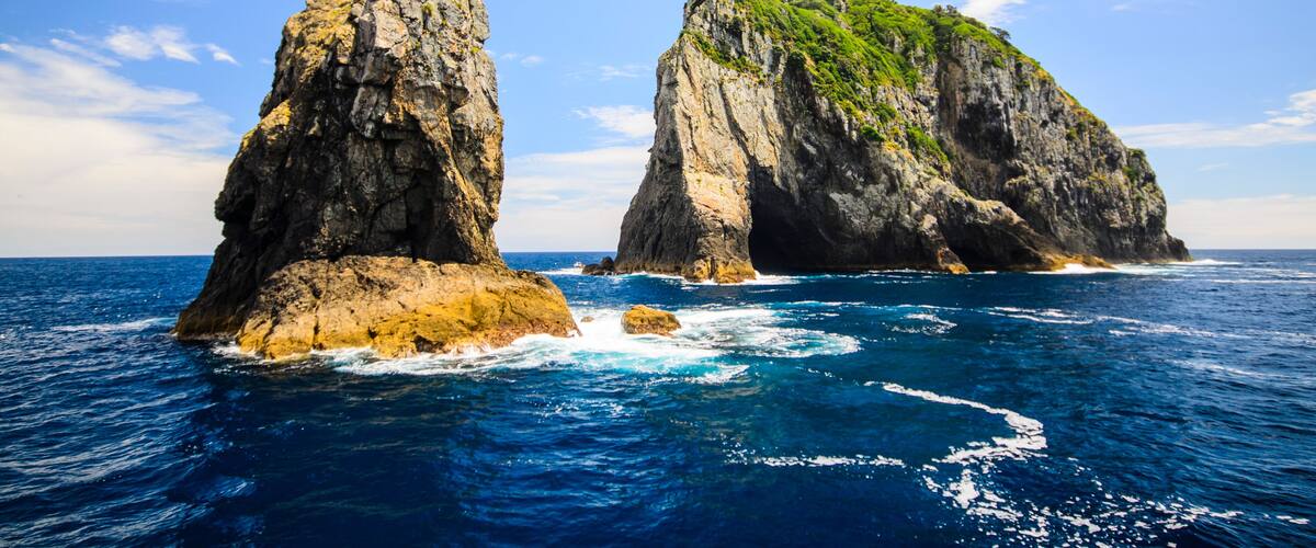 Two prominent rock formations in the ocean with clear blue water and a partly cloudy sky.