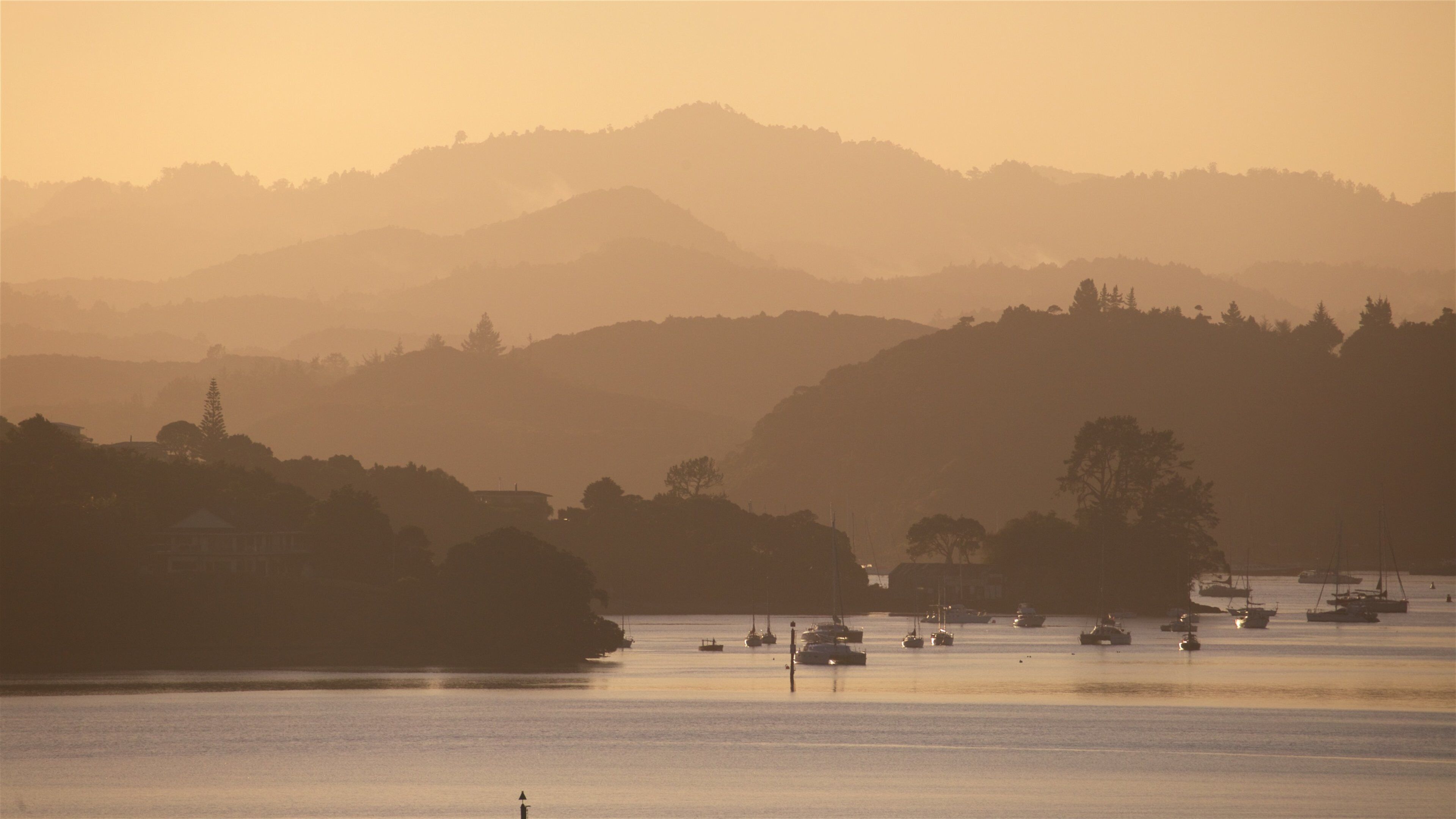 A coastal landscape with boats on calm waters, hills in the background, and a hazy sky.