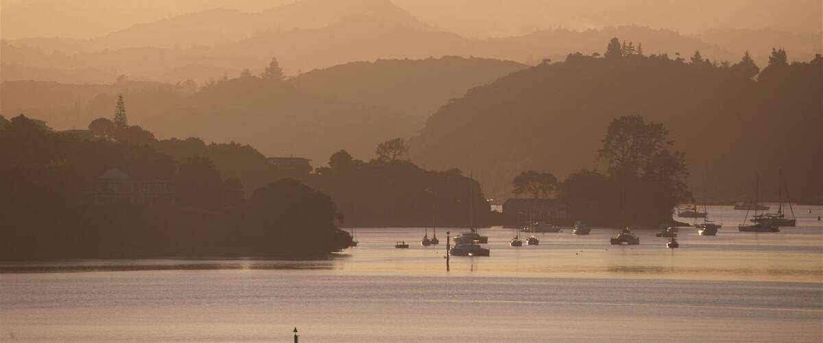 A coastal landscape with boats on calm waters, hills in the background, and a hazy sky.