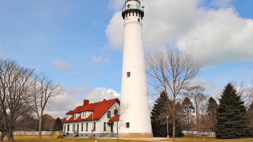 A white lighthouse with a red roof, a white building with a red roof, and a white fence in a grassy area with trees.