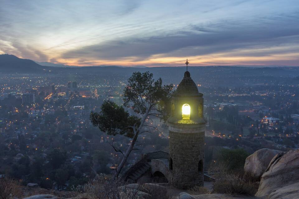Une tour surmontée d’une lanterne illuminée, visible au crépuscule, qui domine un paysage urbain avec des montagnes lointaines.