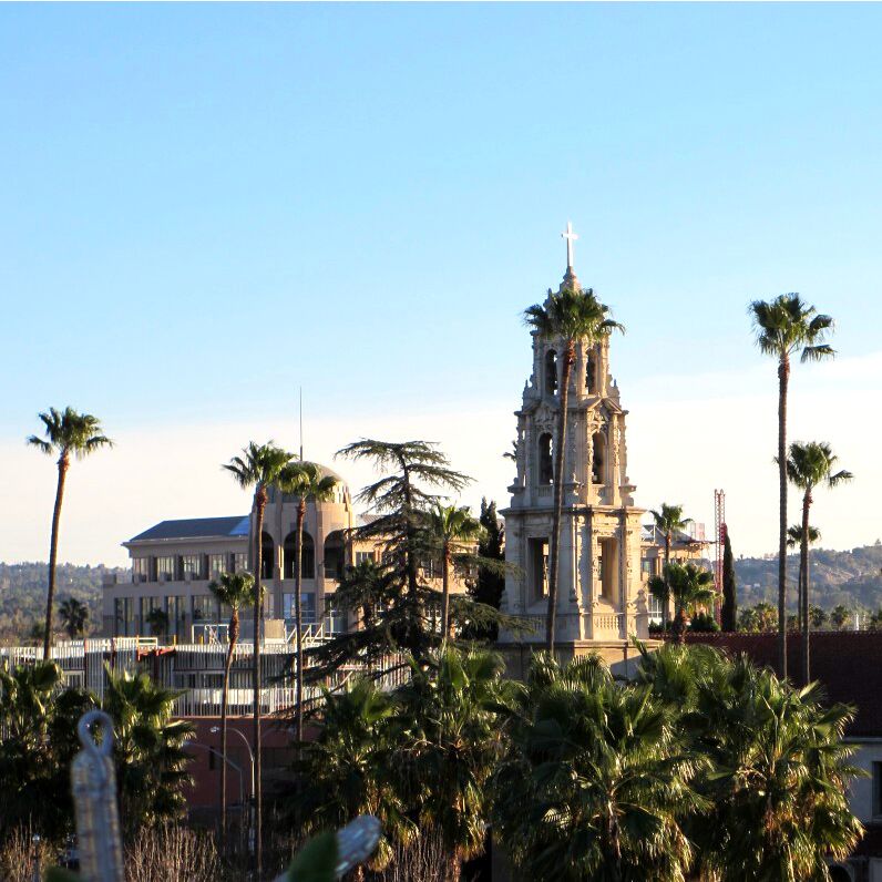 A church with a bell tower, palm trees, and a building with a dome in the background.