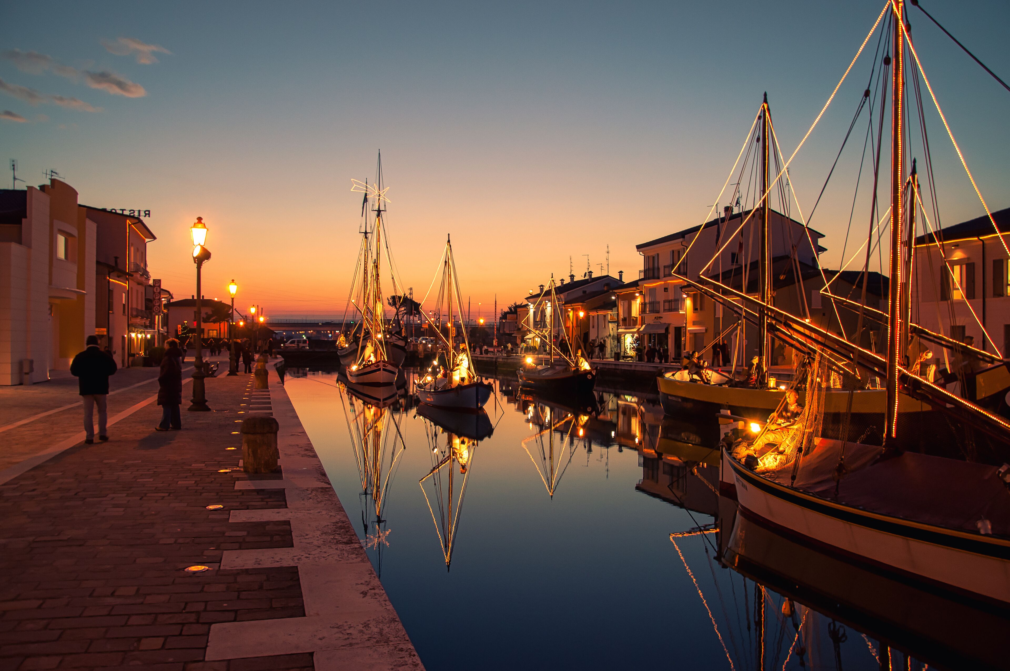 Un paseo marítimo al anochecer, con barcos amarrados, una superficie acuática tranquila que refleja las luces, y edificios a lo largo del malecón.