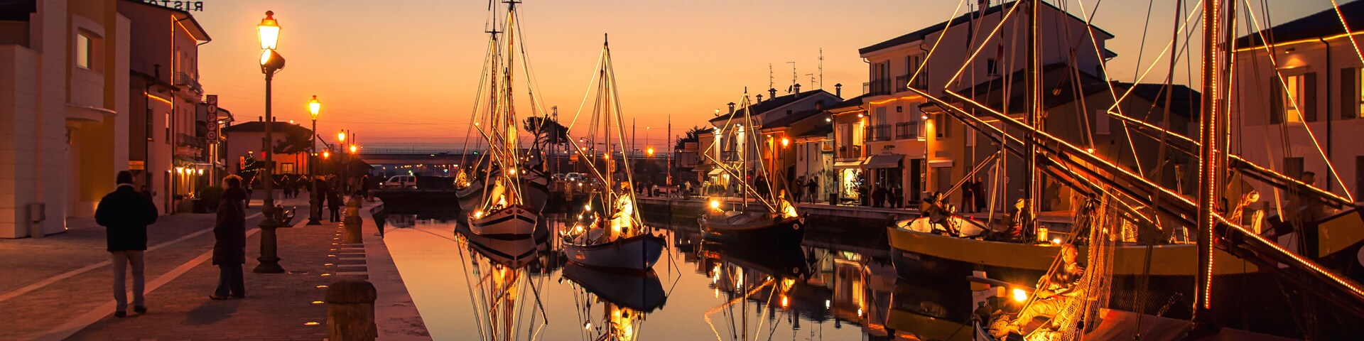 Un paseo marítimo al anochecer, con barcos amarrados, una superficie acuática tranquila que refleja las luces, y edificios a lo largo del malecón.