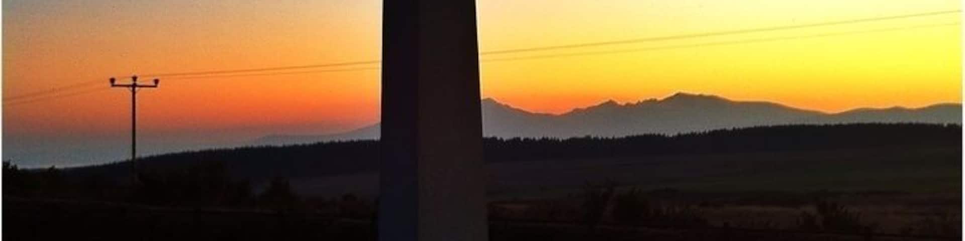 A cross monument with inscriptions, set against a sunset sky with distant hills.