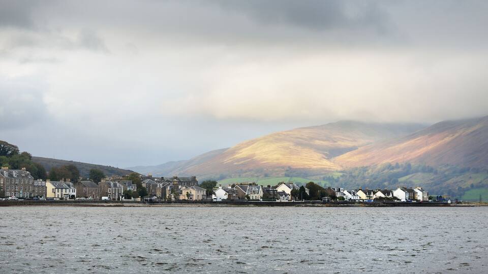 A coastal town with buildings along the shore, a body of water in the foreground, and mountains in the background.