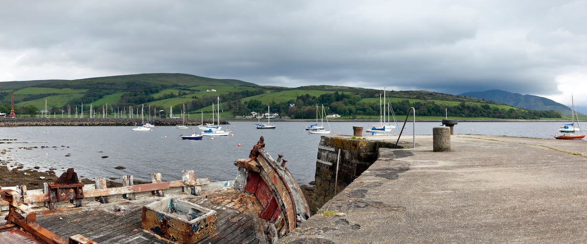 A harbor with boats, a concrete dock, and green hills in the background.
