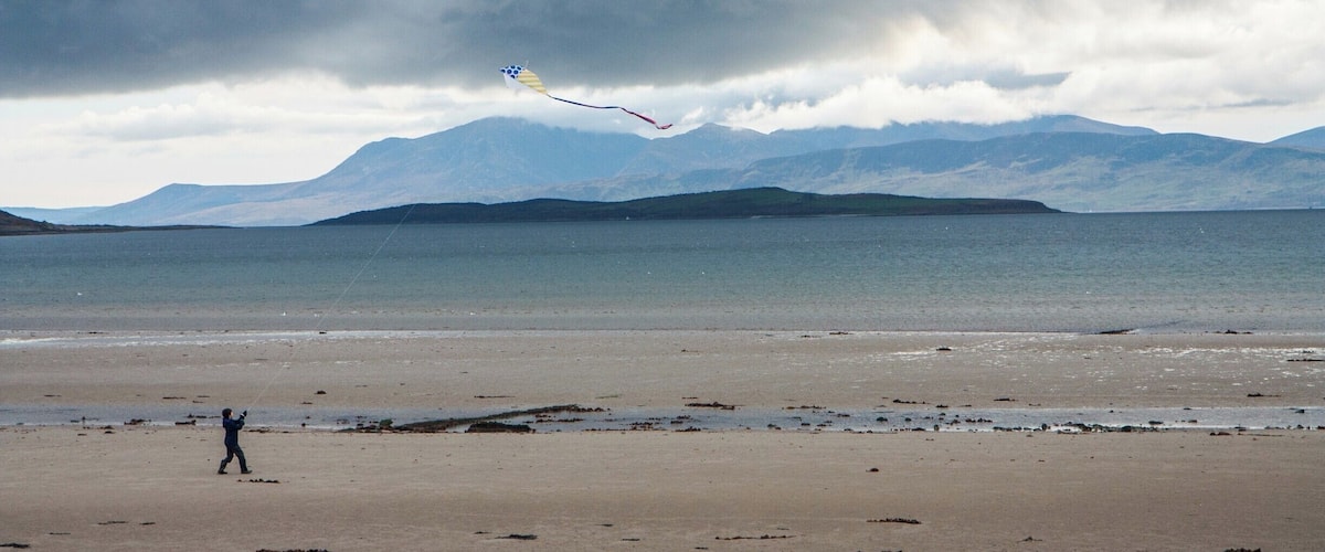 A beach with a person flying a kite, mountains in the background, and a cloudy sky.