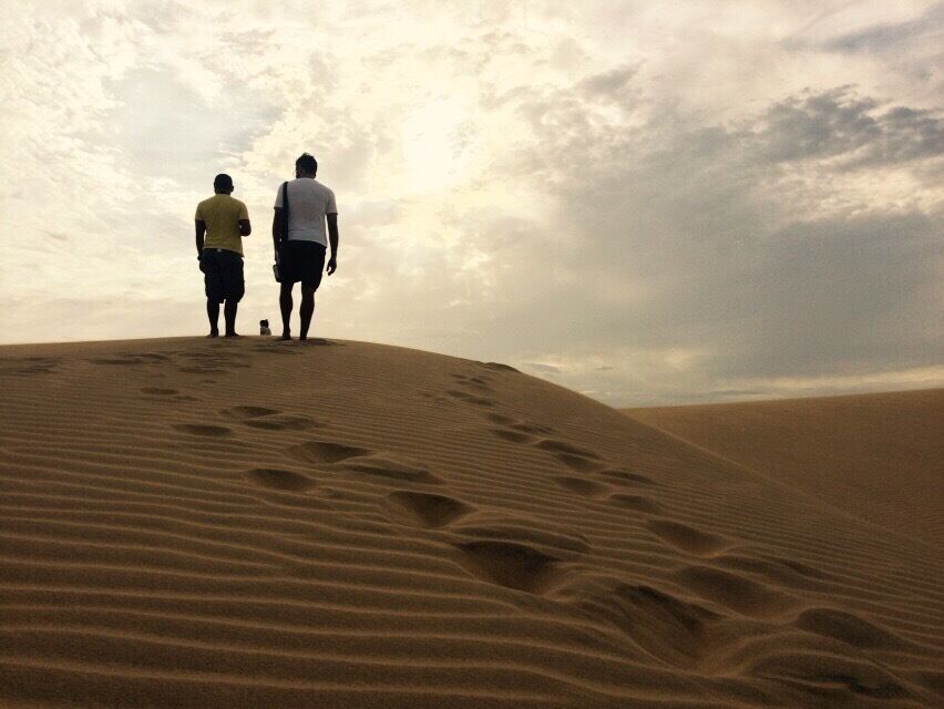 Two people walking on a sandy dune under a cloudy sky.