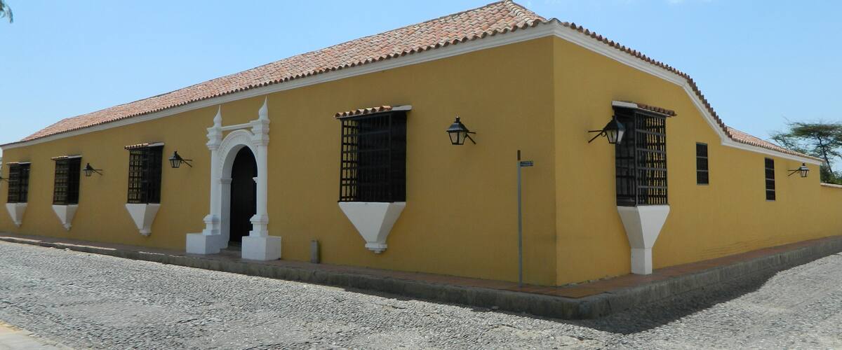 Un edificio amarillo con techo de tejas rojas, detalles decorativos blancos y rejas de ventana negras.