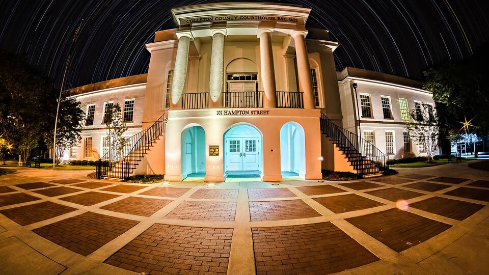 The Colleton County Courthouse at night with illuminated blue doors and columns, surrounded by a paved courtyard.