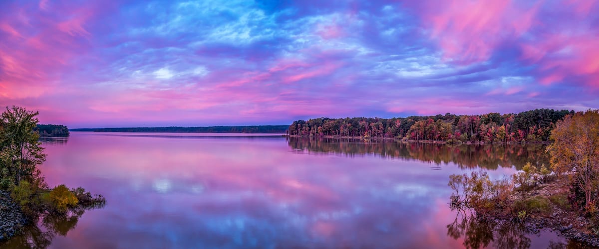 A calm lake with a reflection of the sky, surrounded by trees with autumn foliage.
