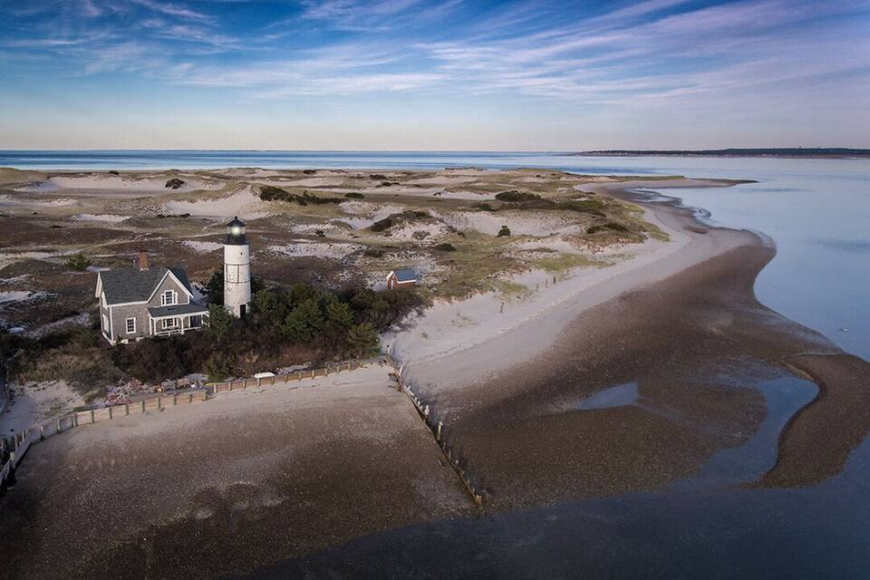 A coastal landscape with a lighthouse, sandy beach, and a house near the water.