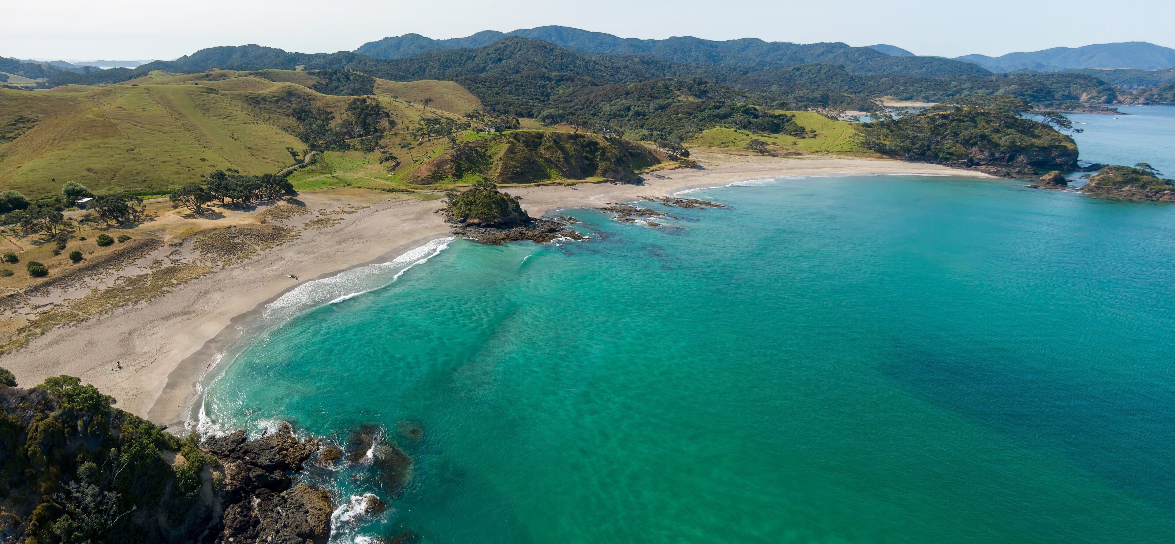 Paisaje costero con playa, aguas turquesas cristalinas y colinas onduladas al fondo.
