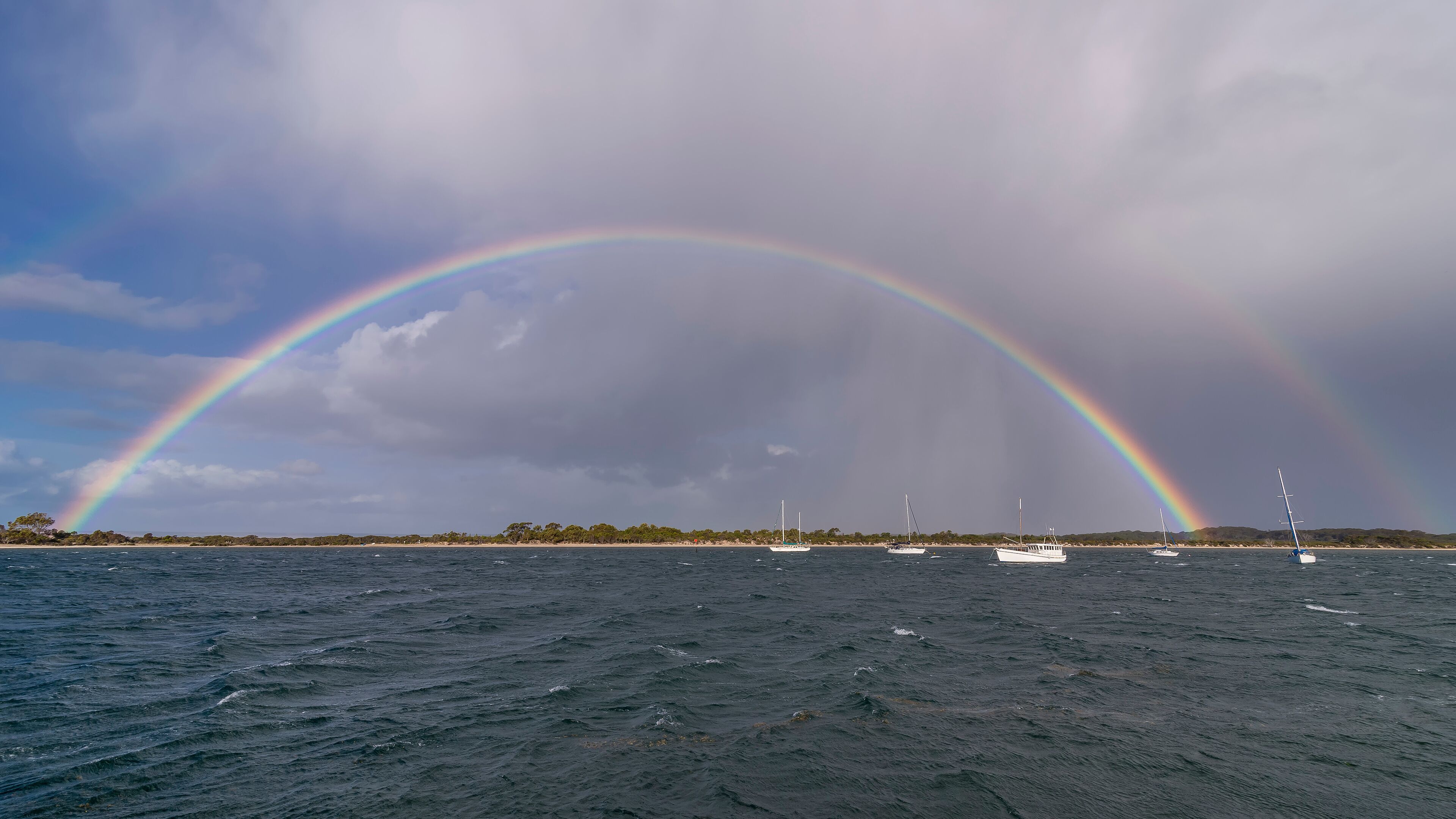 A double rainbow over a body of water with boats and a distant shoreline.