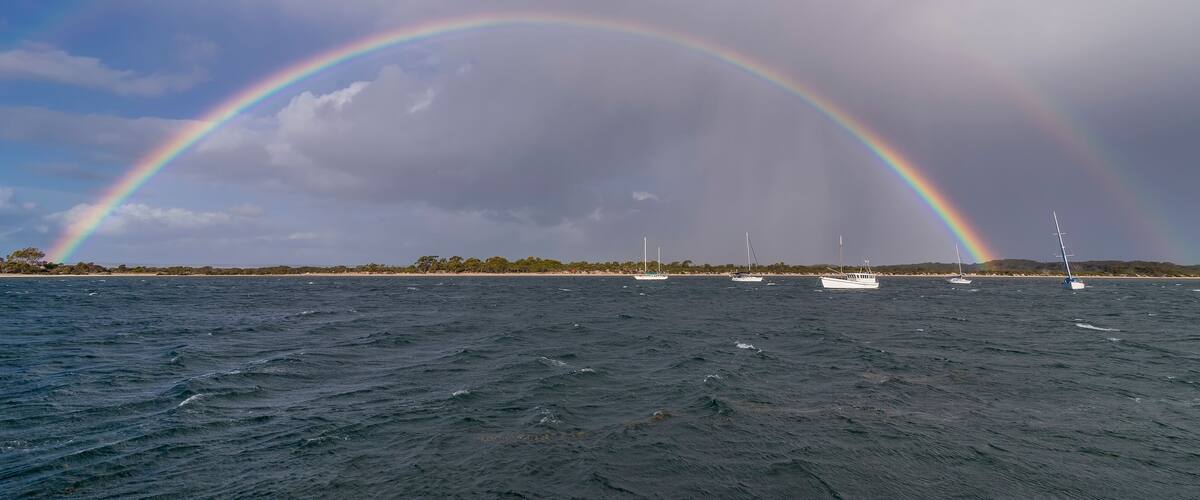 A double rainbow over a body of water with boats and a distant shoreline.