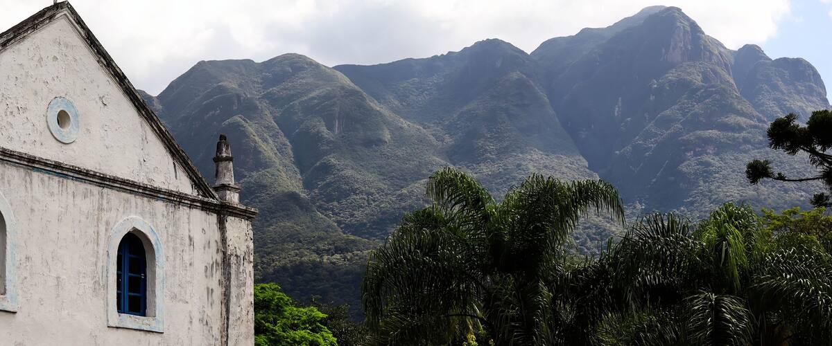 A white church with blue windows and a bell tower, surrounded by lush greenery and mountains in the background.