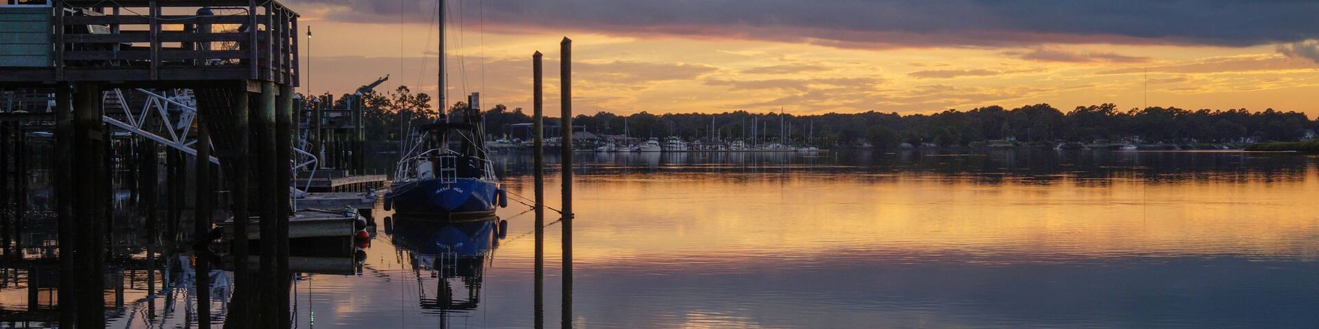 A calm lake at sunset with boats docked at a pier, surrounded by trees and a cloudy sky.