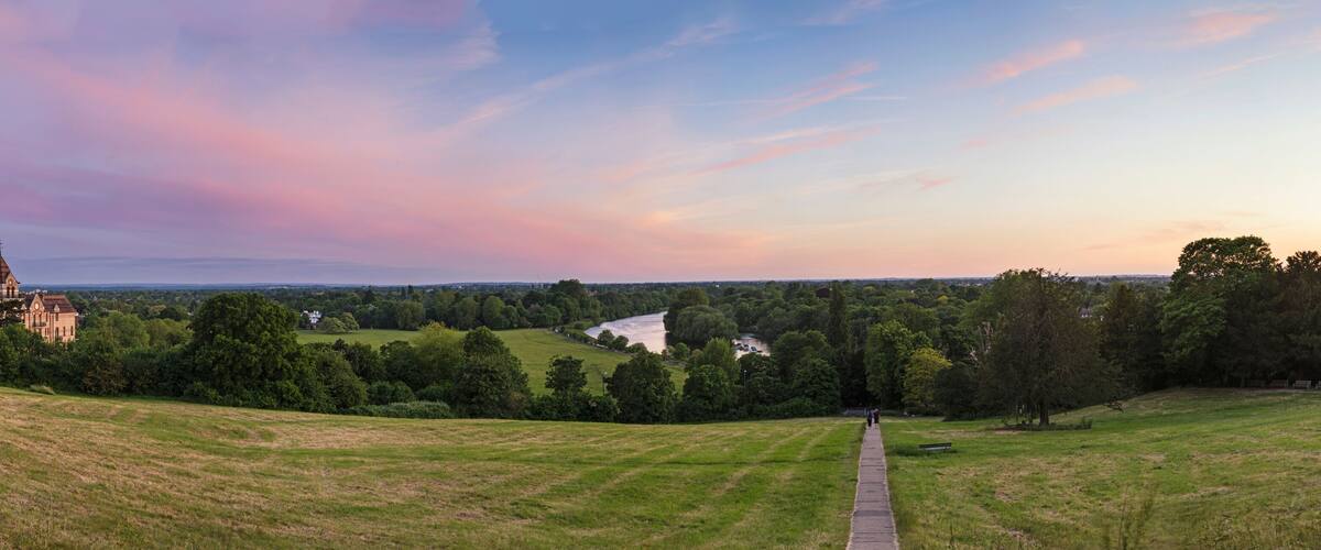 A grassy hill with a pathway leading to a historic building and a river in the distance.