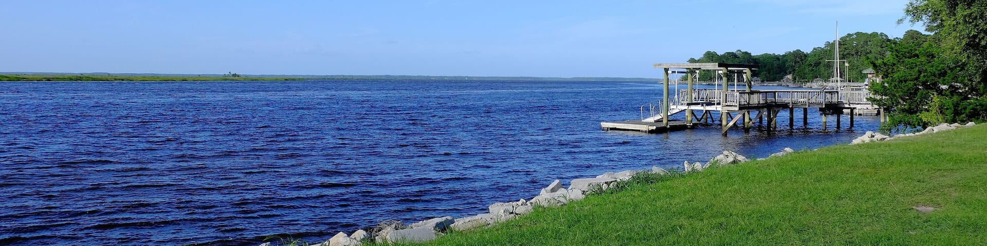 A calm lake with a wooden pier extending into the water, surrounded by green grass and trees.
