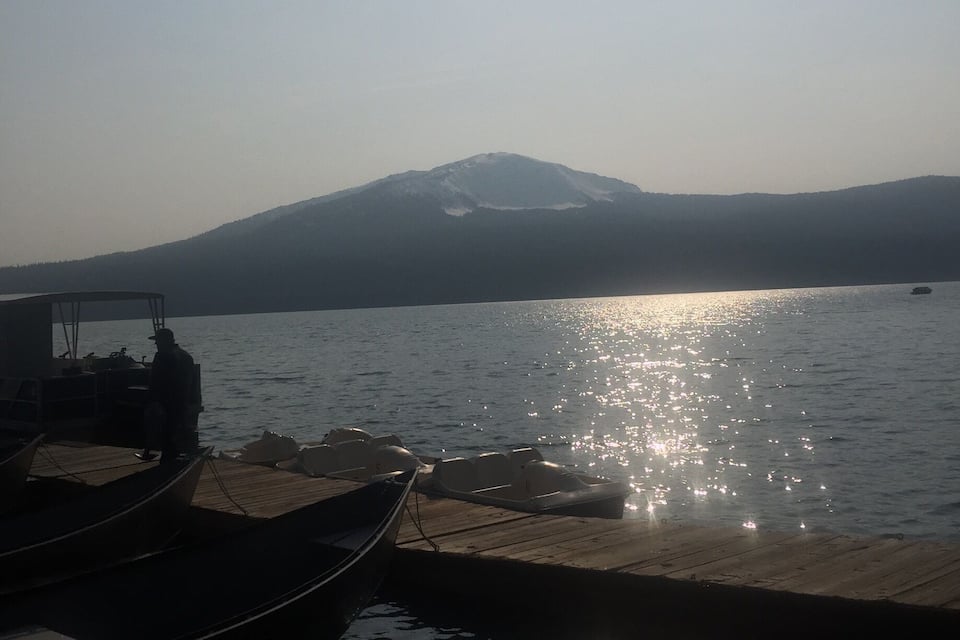 A calm lake with boats docked at the pier, a mountain in the background, and the sun reflecting on the water's surface.