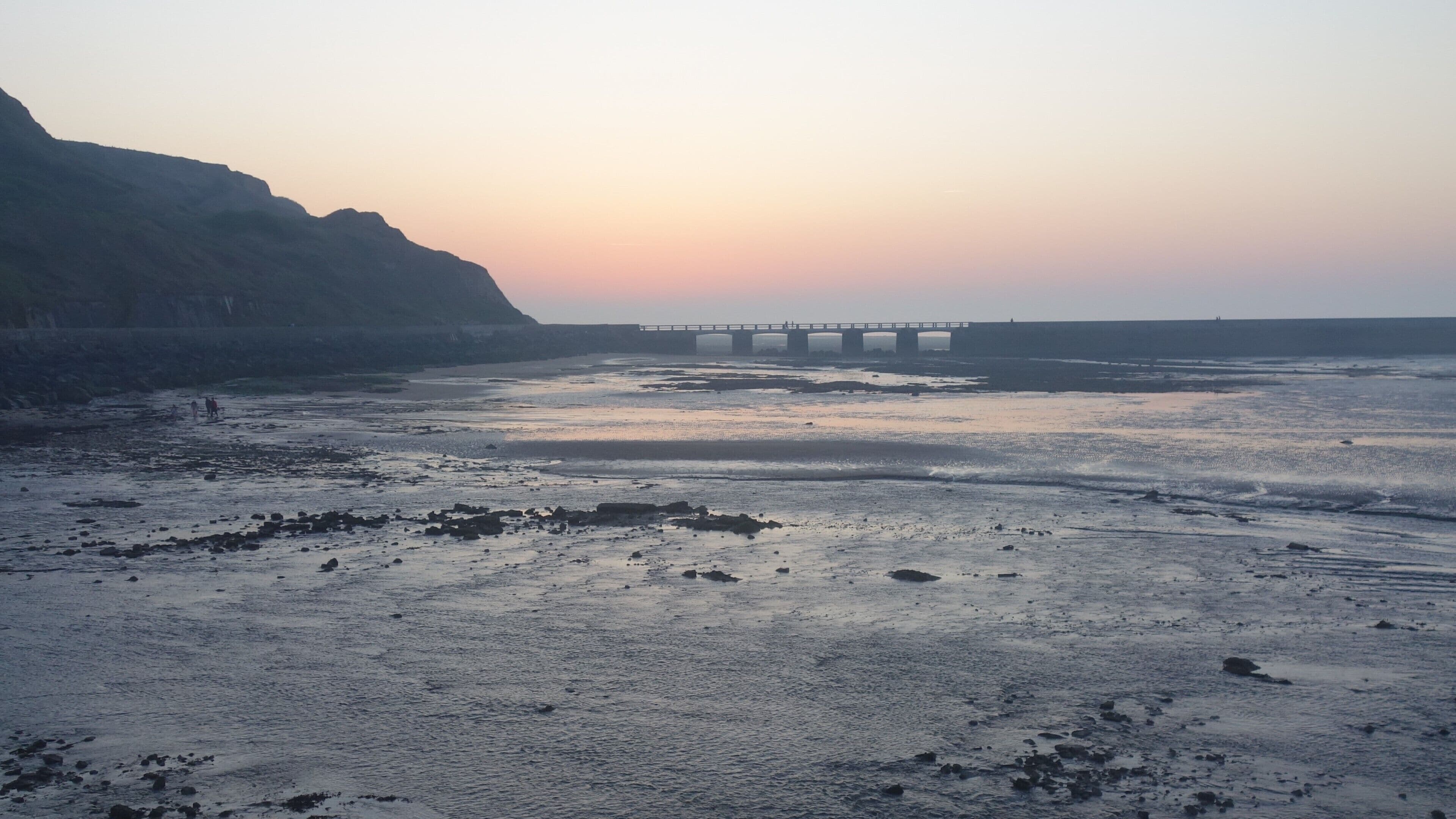 A coastal landscape with a rocky beach, calm sea, and a bridge in the distance.