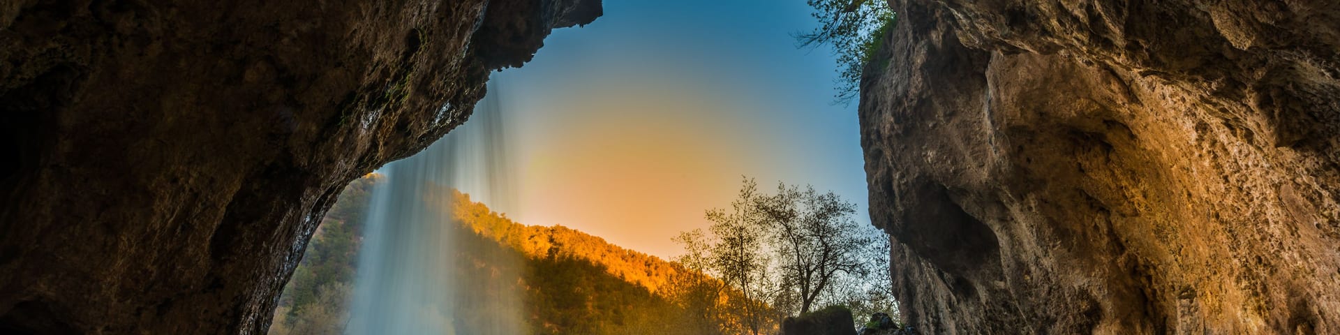 A cave opening with a waterfall inside, surrounded by trees and rocky terrain.