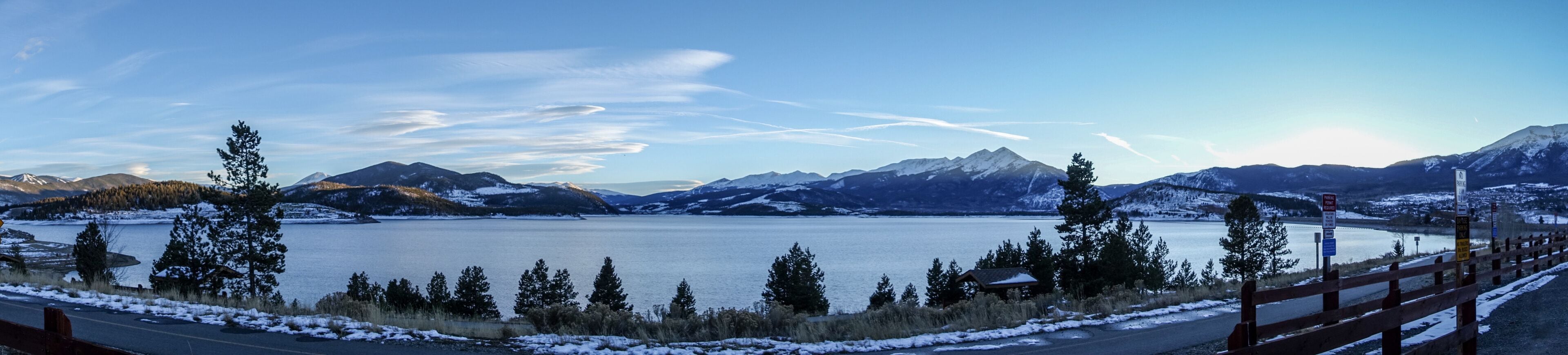 A snow-covered lake with mountains in the background and a wooden fence in the foreground.