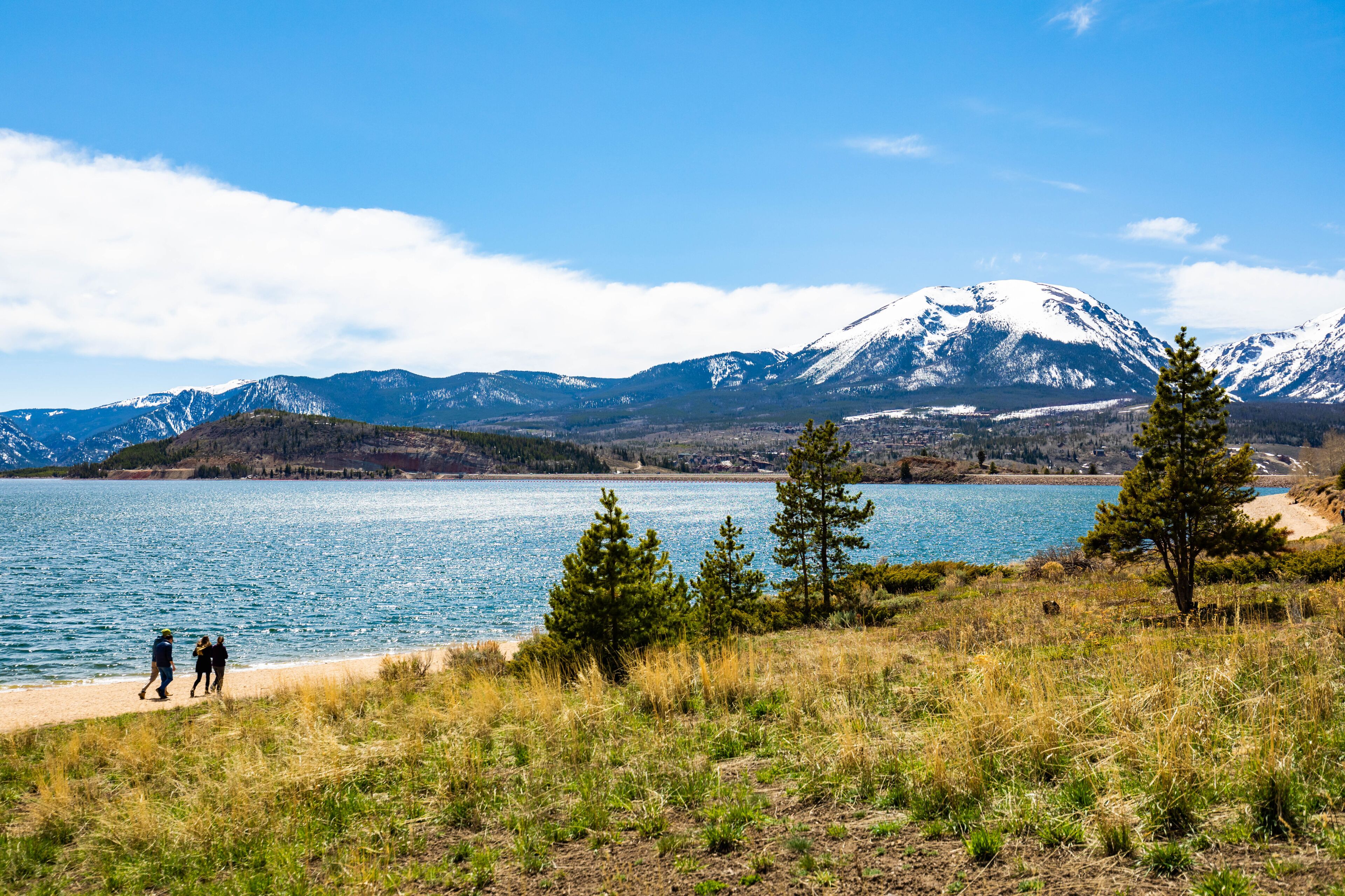 A lake with mountains in the background, a clear sky, and a few people walking along the shore.
