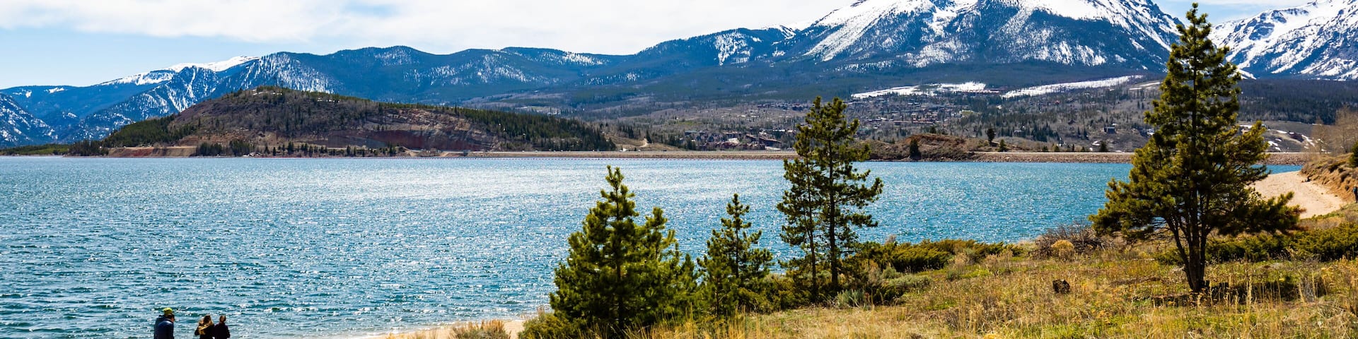 A lake with mountains in the background, a clear sky, and a few people walking along the shore.
