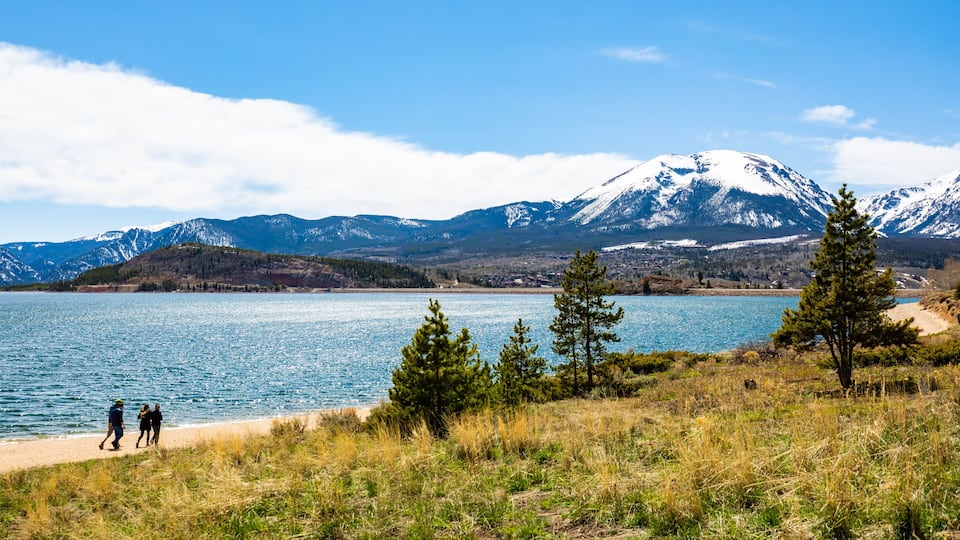 A lake with mountains in the background, a clear sky, and a few people walking along the shore.