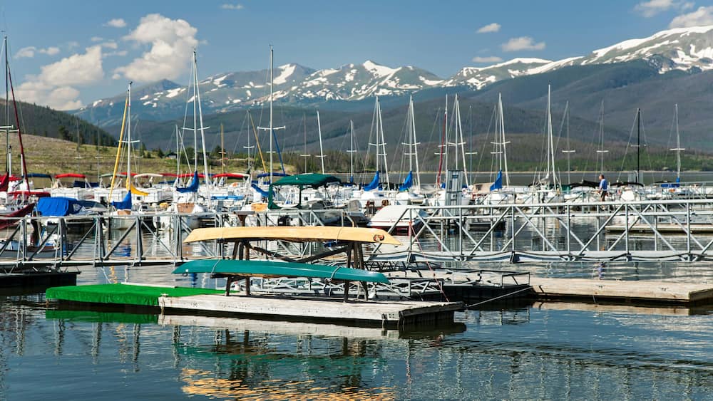 Un port de plaisance où sont amarrés des voiliers, une jetée surmontée d’une tonnelle verdoyante, et des montagnes enneigées en arrière-plan.
