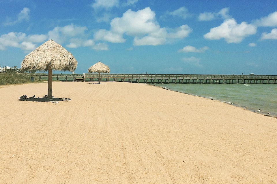 A sandy beach with two thatched umbrellas and a wooden pier extending into the water.