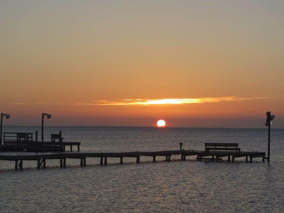 A sunset over a calm sea with a pier extending into the water, featuring benches and street lamps.