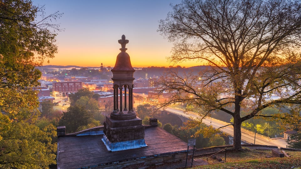 A sunset view of a cityscape with a prominent monument on a rooftop, surrounded by trees and a bridge in the background.