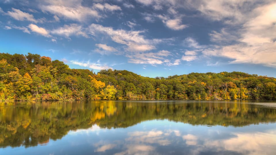 A calm lake reflecting the autumn foliage and a clear sky.