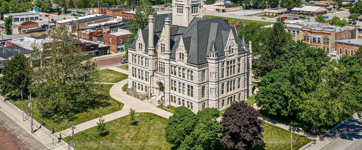 An aerial view of a historic building with a clock tower, surrounded by greenery and urban structures.
